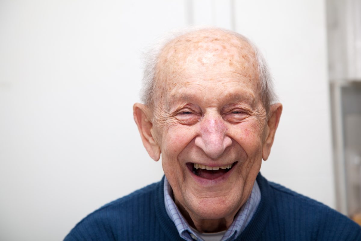 Senior adult male portrait; he is 90 years old. In front of his out of focus white kitchen wall.